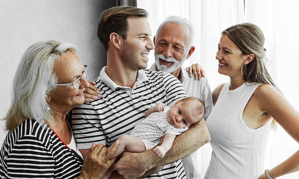 three generations of family members smiling together while holding newborn baby