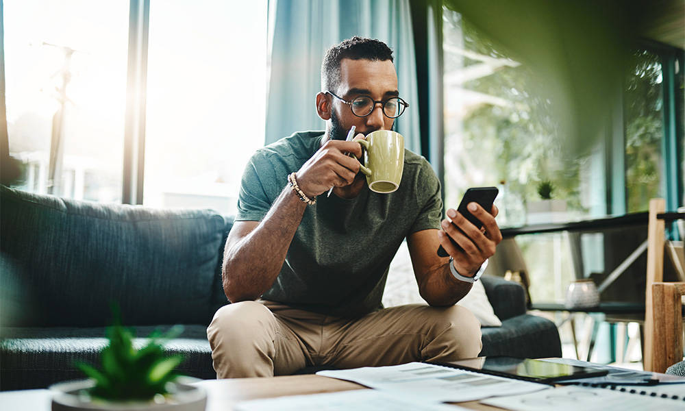 Young man using a smartphone and drinking coffee while going over his finances in his living room at home.