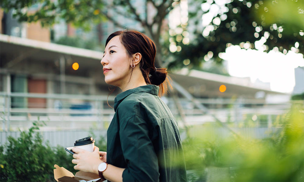 Smiling young businesswoman using smartphone while having a healthy salad lunch box with a cup of coffee outdoors in an urban park.