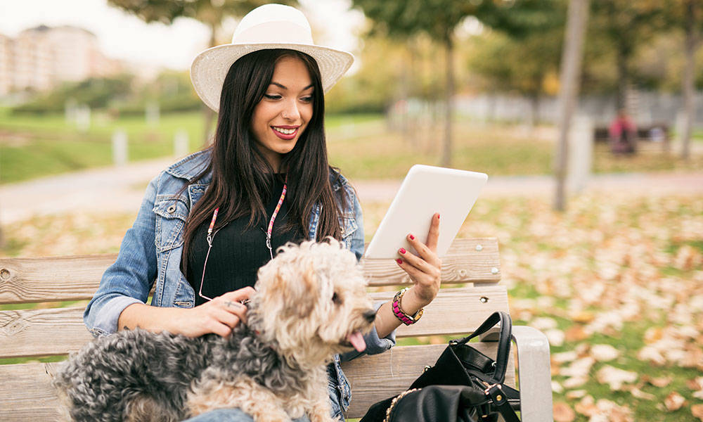 woman in denim jacket and hat looking at tablet with her dog on park bench
