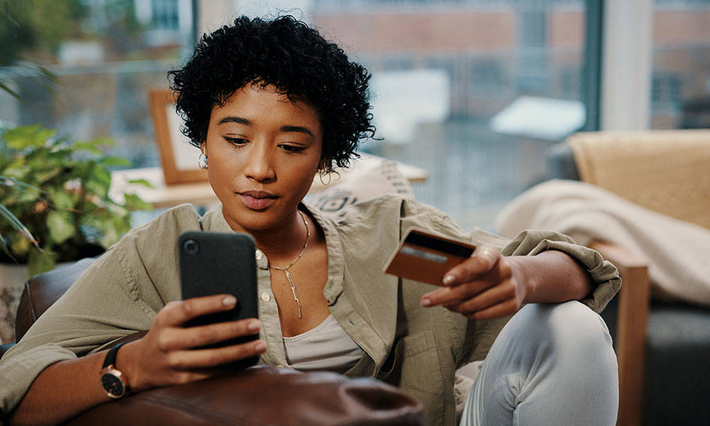 young woman holding her credit card while using her smartphone at home