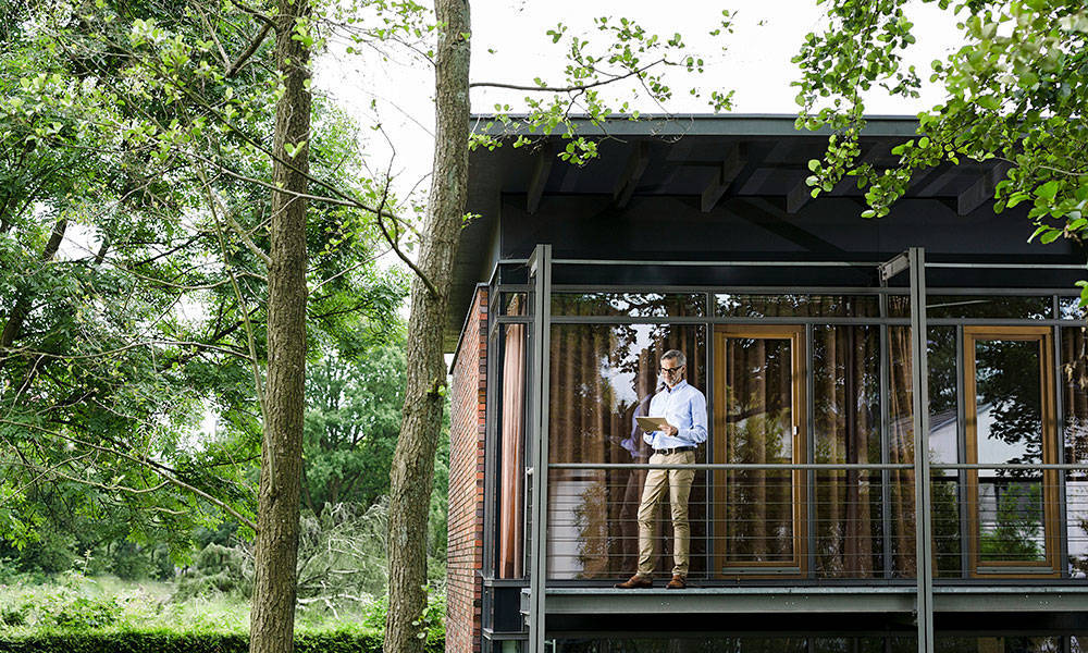 man standing outside of his house on tablet, surrounded by trees