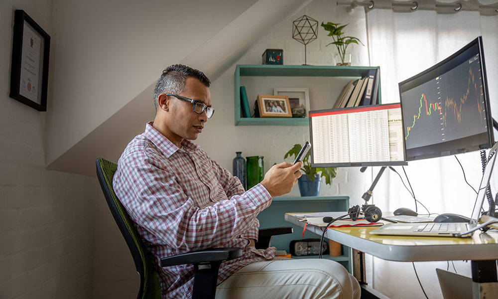 man looking at his smartphone and evaluating market conditions on desktop computer