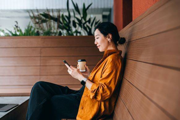 Woman enjoying coffee while reading smartphone outdoors.
