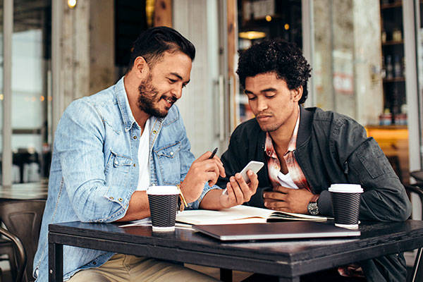 Two friends sitting together at coffee shop using Popmoney on their smartphones to pay each other.