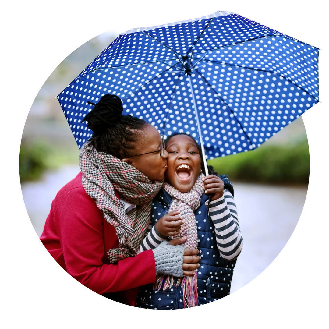 Mother kissing her daughter, which makes her laugh, under an umbrella.