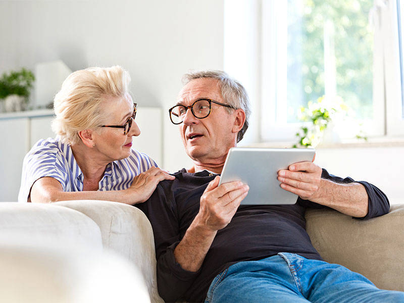 Elderly man sitting on sofa in the living room at home and holding digital tablet in hands, talking with his wife.