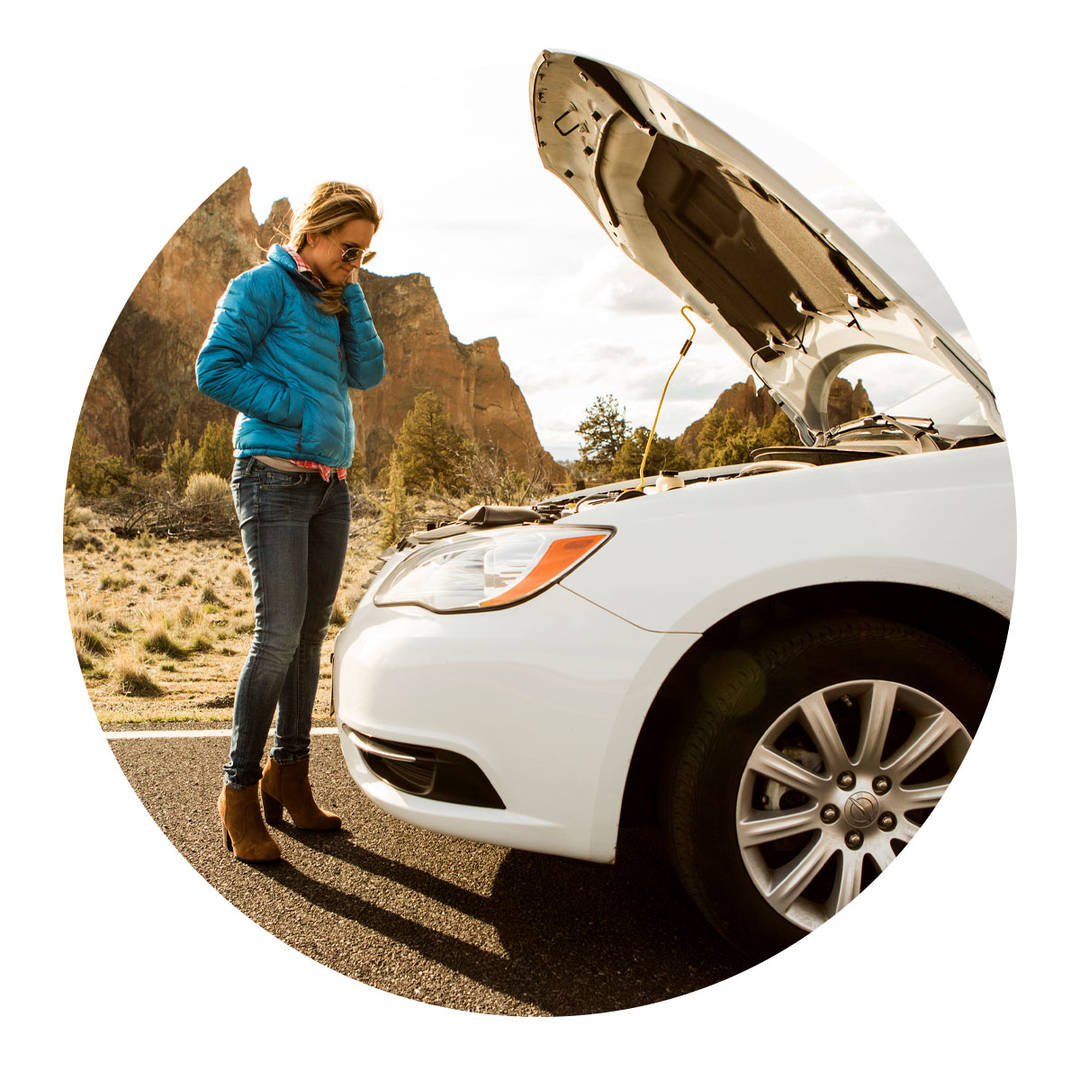 A woman calling for assistance while looking down at the broke down car's engine in the desert.