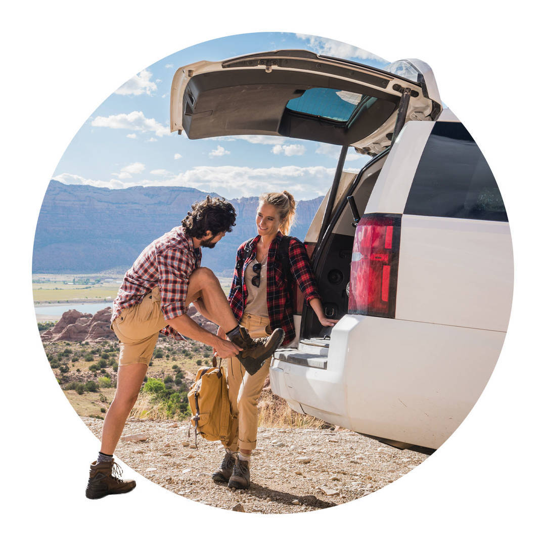 At a scene lookout location in the mountains, a young couple tying shoes under the white SUV's liftgate.