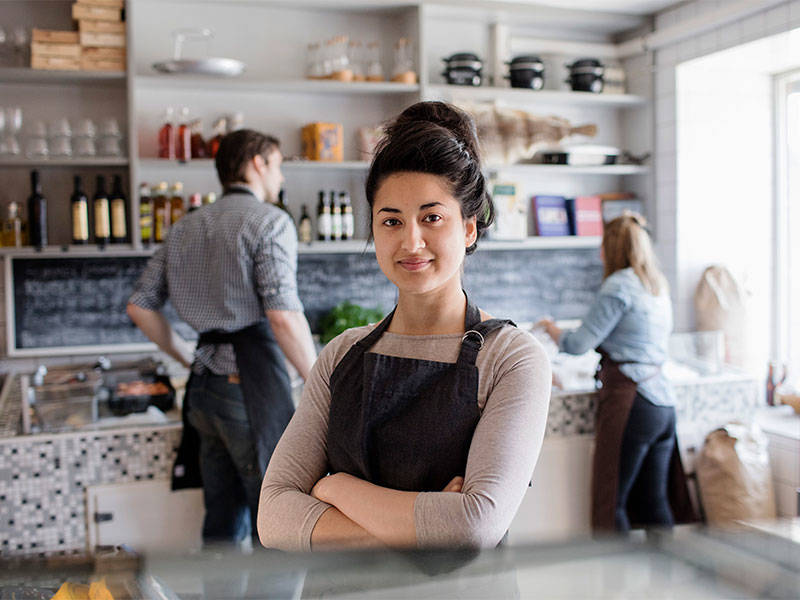 Confident small business owner standing at the restaurant counter while her two employees are diligently working behind.