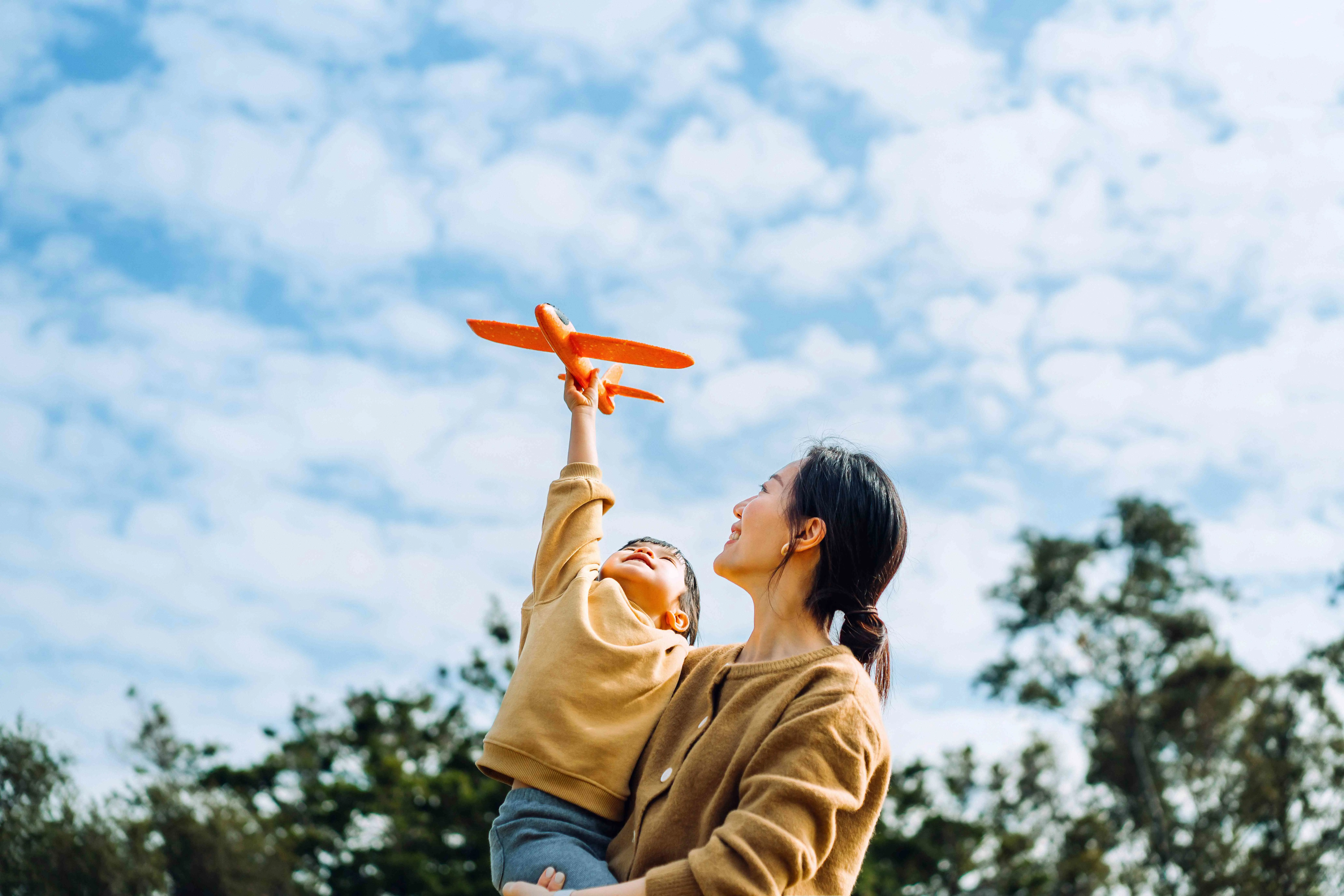 Young Asian mother and lovely little daughter spending time together outdoors, playing with airplane toy and smiling joyfully in park on a lovely sunny day against beautiful blue sky